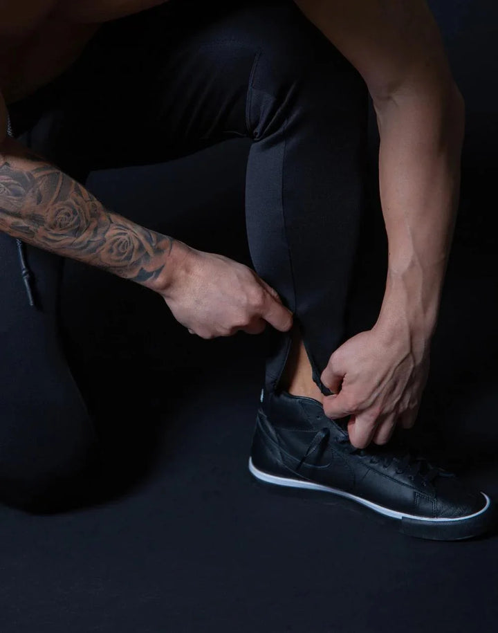 Man wearing black slim joggers with zipper cuffs, adjusting pants over black sneakers