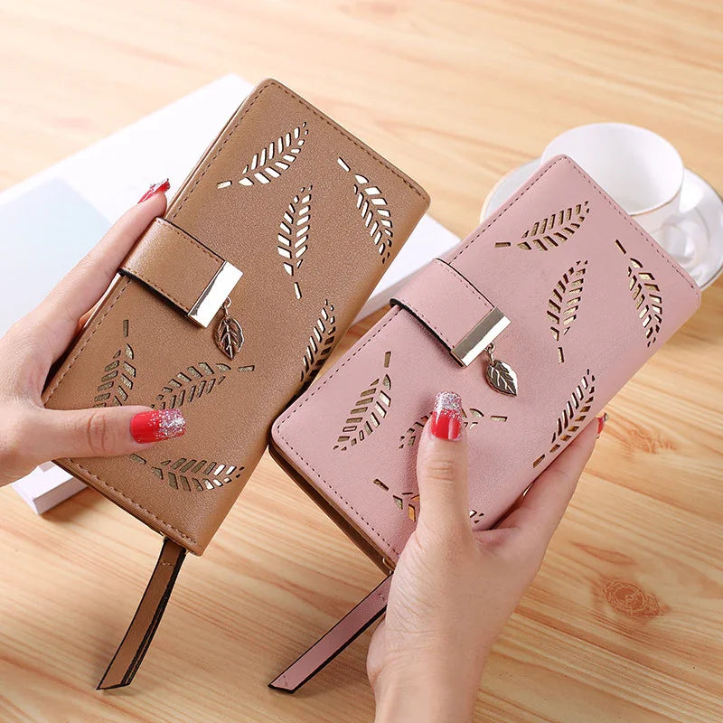 Two women's wallets with leaf cutout design, one brown and one pink, held by hands with red glitter nails on wooden table