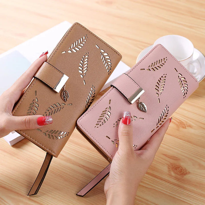 Two women's wallets with leaf cutout design, one brown and one pink, held by hands with red glitter nails on wooden table