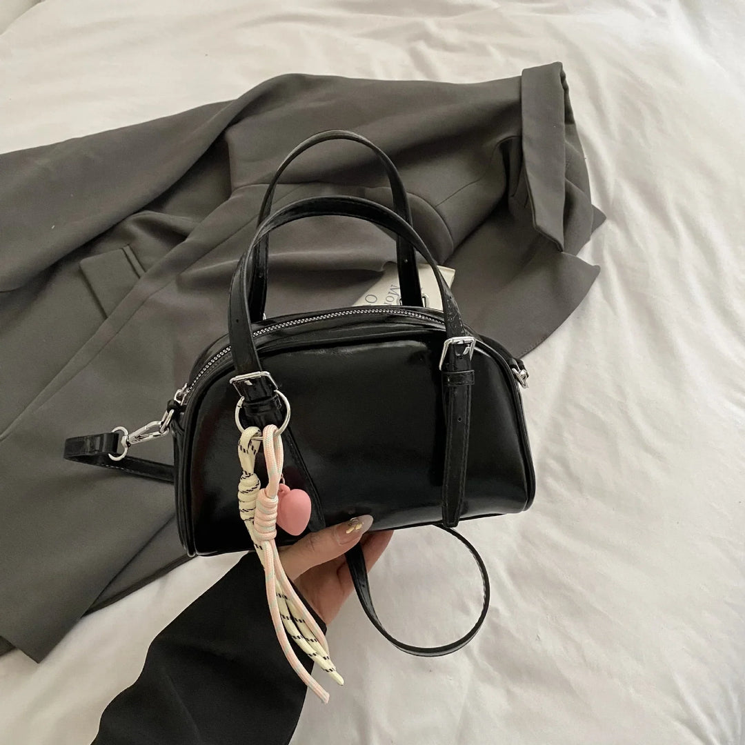 Glossy black mini handbag with double handles and pink heart keychain, held above gray blazer on white bed