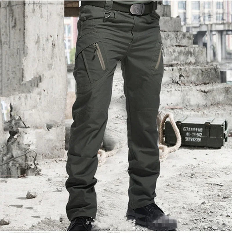 Men's green tactical cargo pants with multiple zippered pockets worn in urban industrial setting