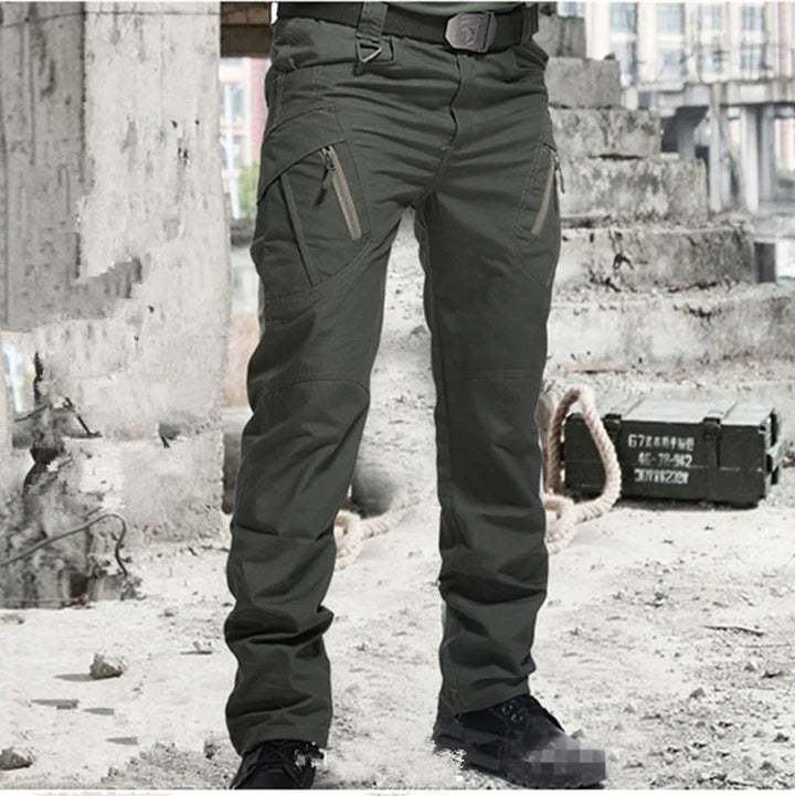 Men's green tactical cargo pants with multiple zippered pockets worn in urban industrial setting