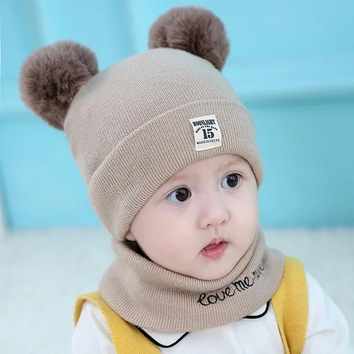 Cute baby wearing beige knit hat with pom-pom ears and matching neck warmer, blue background