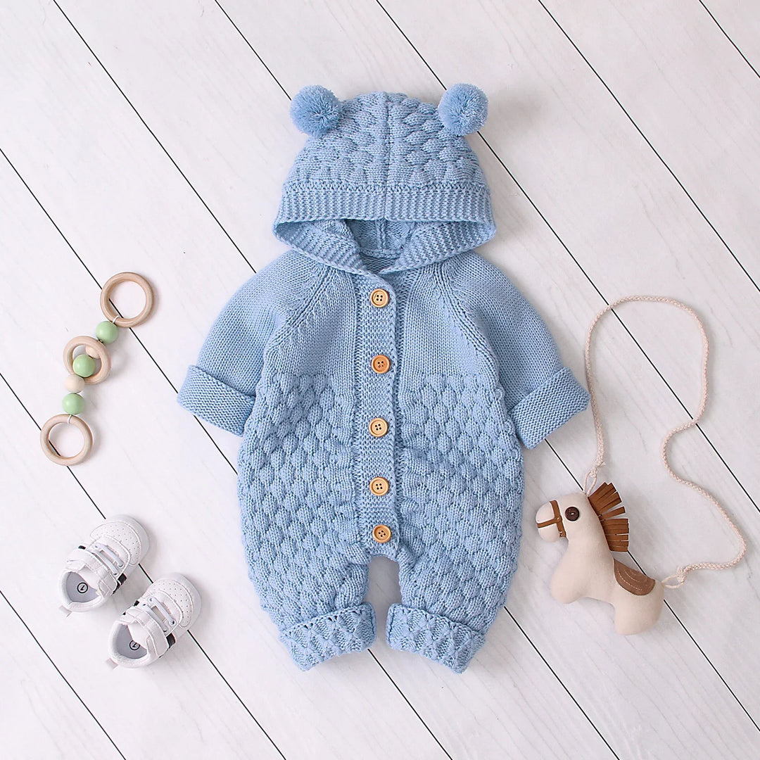 Blue knitted baby romper with hood and pom-poms, wooden buttons, baby shoes, toy horse, and wooden ring toy on white floor