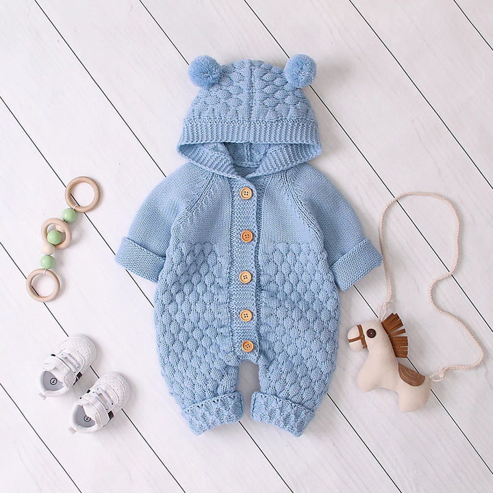 Blue knitted baby romper with hood and pom-poms, wooden buttons, baby shoes, toy horse, and wooden ring toy on white floor