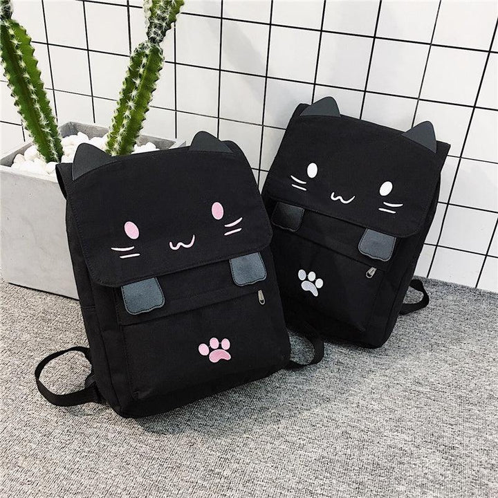 Two black cat-themed backpacks with pink and white facial details and paw prints on gray carpet