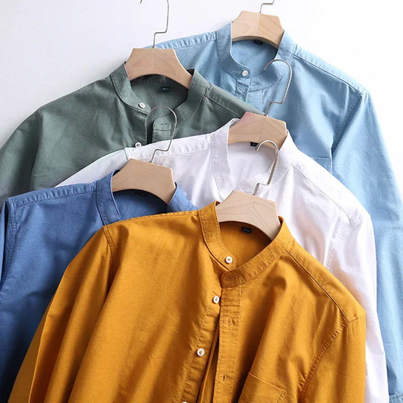 Five mandarin collar shirts in mustard, blue, white, green, and light blue hanging on wooden hangers