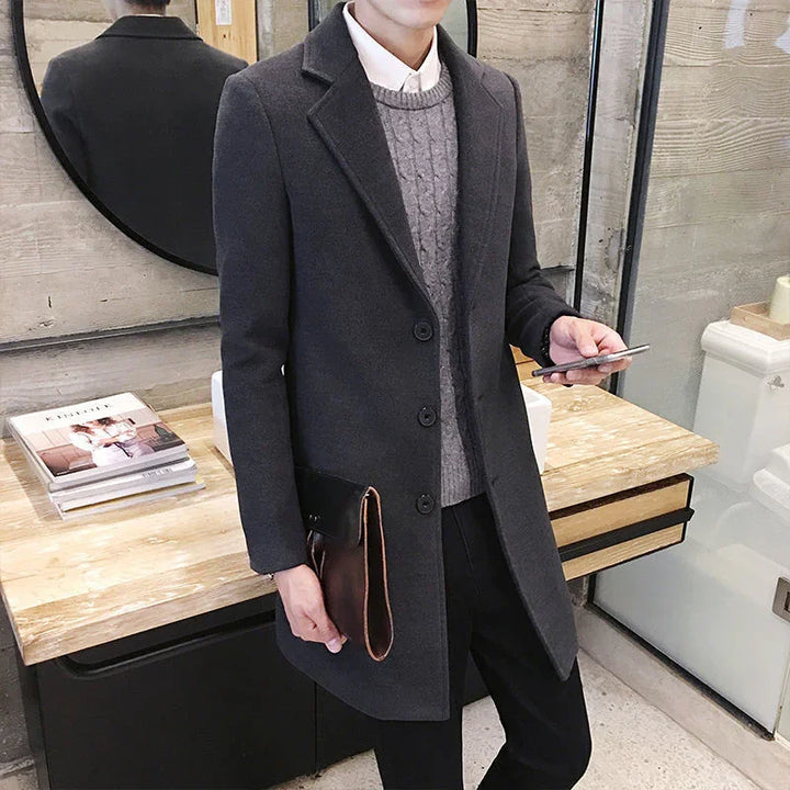 Man wearing dark gray overcoat and gray cable knit sweater holding phone and brown leather clutch indoors