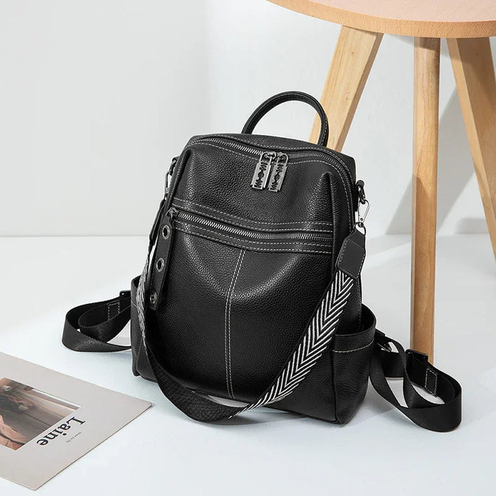 black leather backpack with silver zippers and black-white chevron strap next to wooden chair on white floor