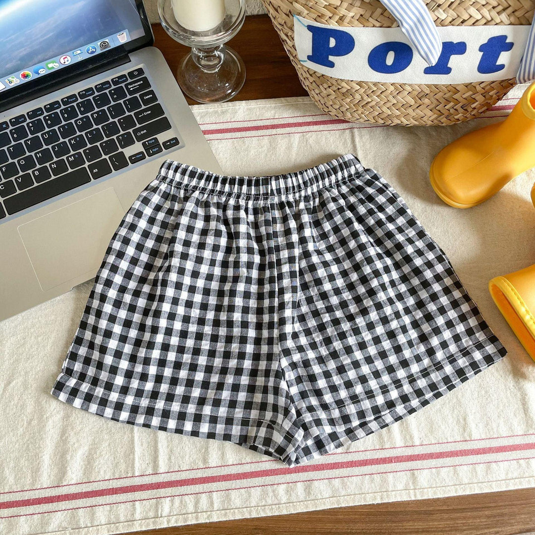Black and white checkered shorts on a table beside a laptop, yellow rain boots, candle, and straw bag