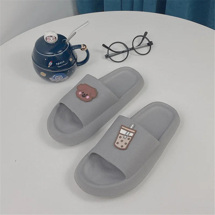 Gray slide slippers with cute dog and bubble tea patches on a white table with black round glasses and a navy space-themed teapot