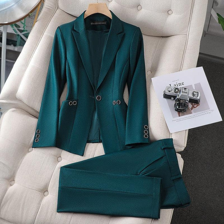 Dark green tailored women's blazer and matching trousers laid on a beige tufted sofa with a vintage camera on paper nearby