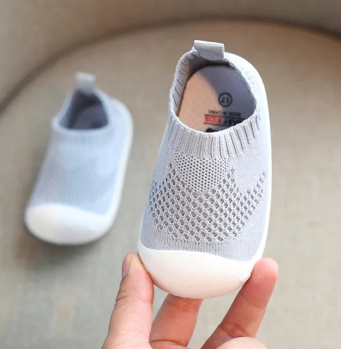 Hand holding gray breathable knit baby shoe with white sole on beige background
