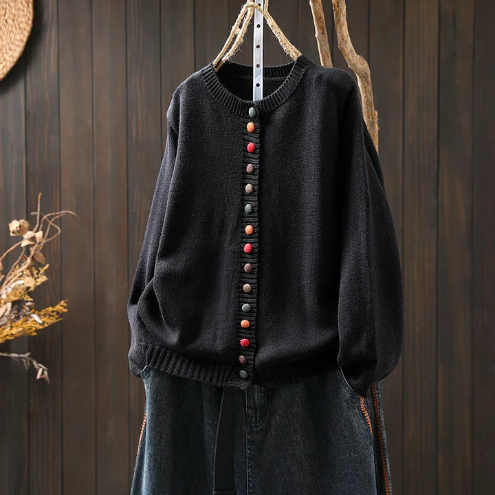 Black knit cardigan with multicolored buttons hanging on rustic rope hanger against wooden backdrop