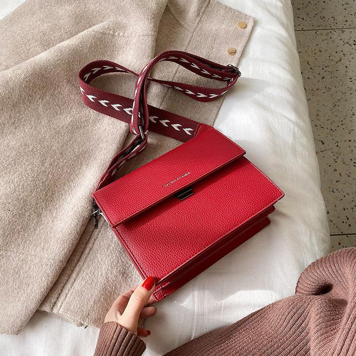 Red leather crossbody bag with patterned strap placed on beige coat and white fabric surface