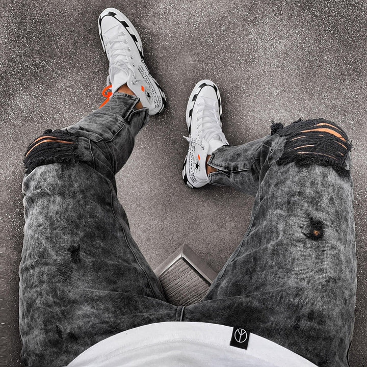 Person wearing distressed black jeans and white sneakers with black and orange accents on gray pavement