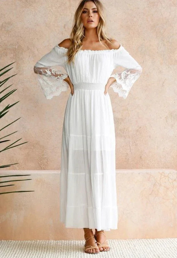 Woman in white off-shoulder maxi dress with lace bell sleeves, standing against beige textured wall