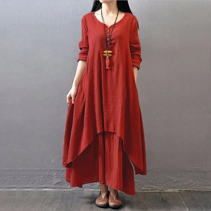 Woman wearing a loose-fit, layered red dress with long sleeves and brown shoes, standing against gray wall