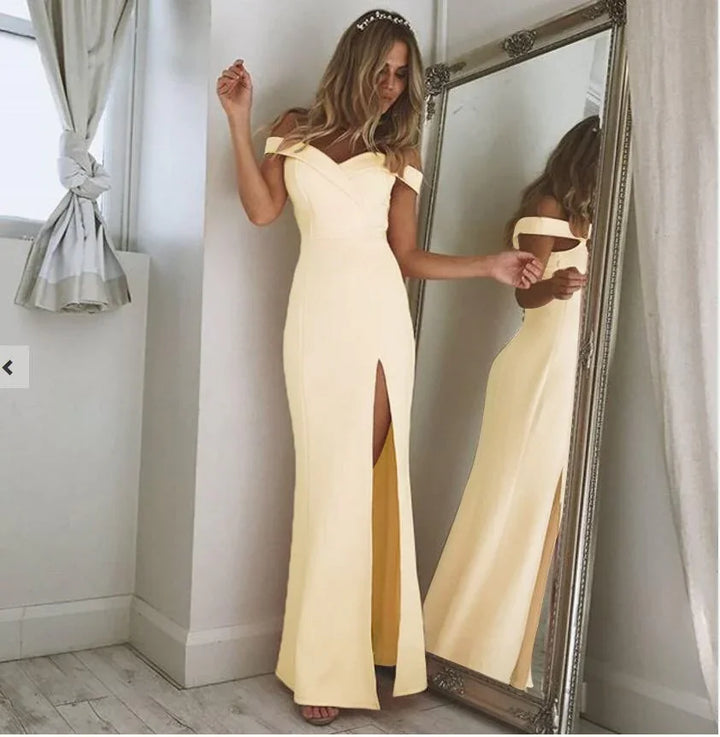 Woman wearing a yellow off-shoulder evening gown with thigh slit, standing beside ornate mirror in minimal room