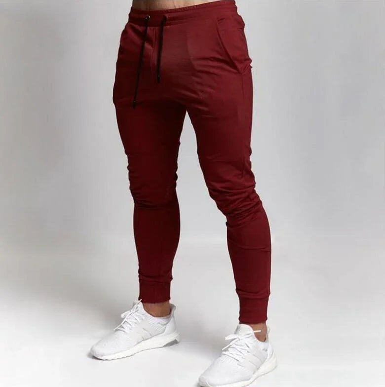 Man wearing slim-fit burgundy jogger pants with black drawstrings and white athletic sneakers on gray background