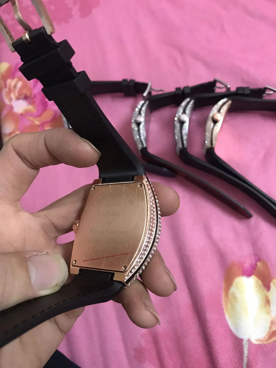 Hand holding rose gold watch with black strap and diamond accents, three similar watches on pink floral background