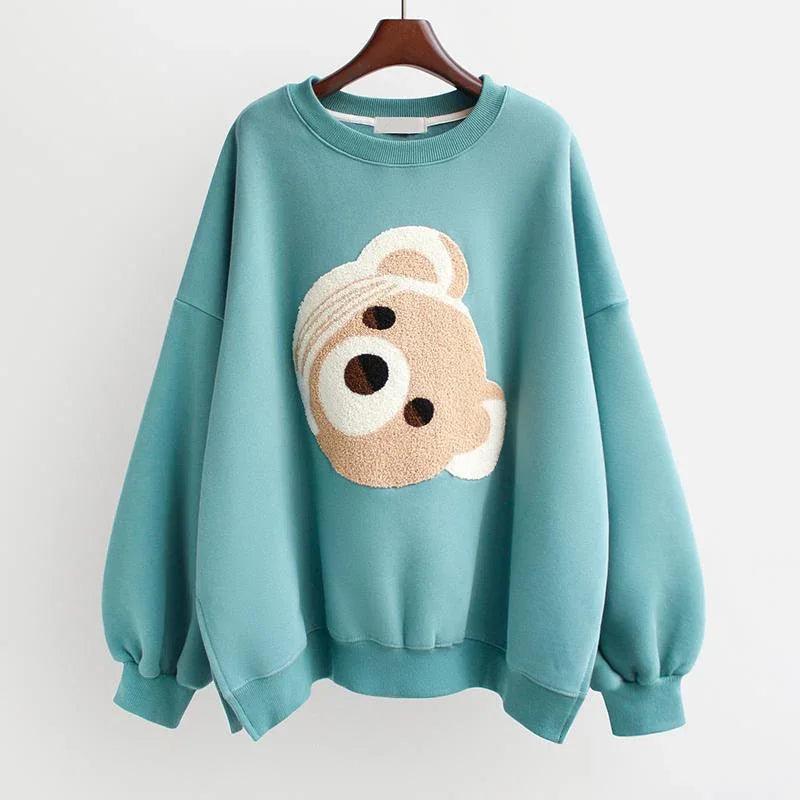 Teal oversized sweatshirt with a textured teddy bear face graphic on the front, hanging on a wooden hanger