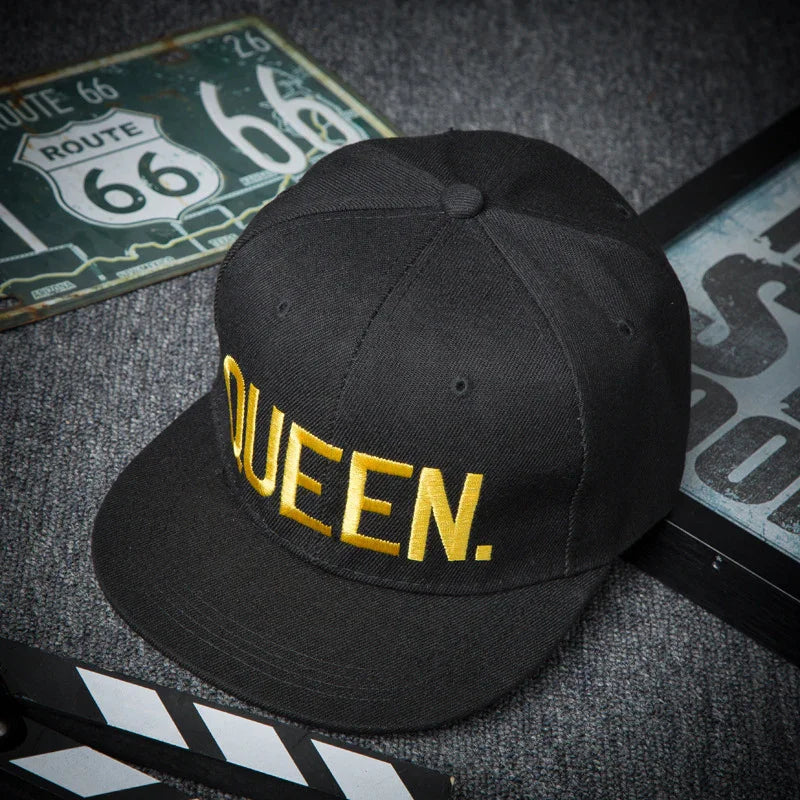Black baseball cap with yellow embroidered QUEEN text, placed on gray carpet near Route 66 signs