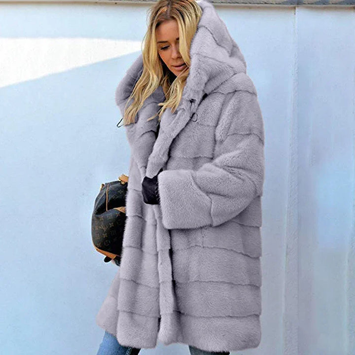Woman wearing a stylish light gray hooded faux fur coat against a blue and white wall