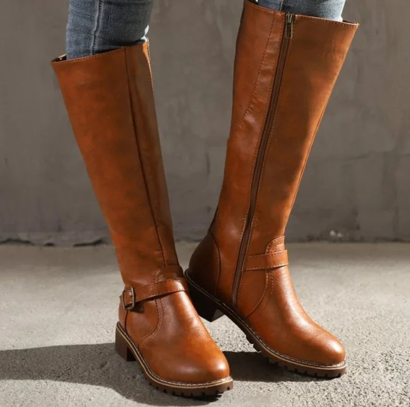 Pair of brown knee-high leather boots with side zippers and buckle detail on concrete floor