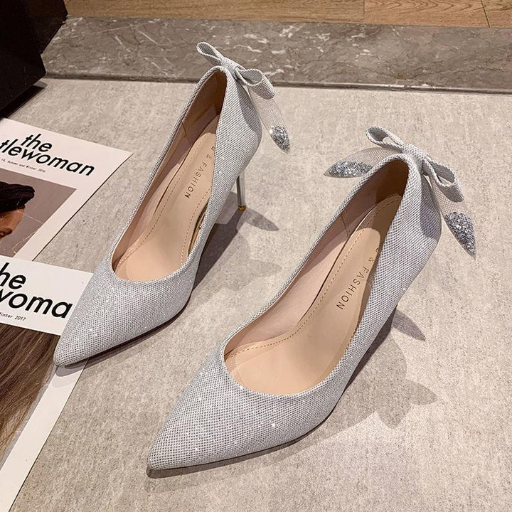 Elegant silver high heel shoes with pointed toes and decorative bows on a stone floor next to a magazine