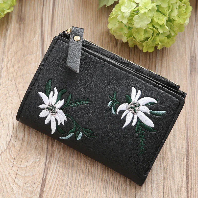 Black leather wallet with white floral embroidery and gold zipper on wooden surface