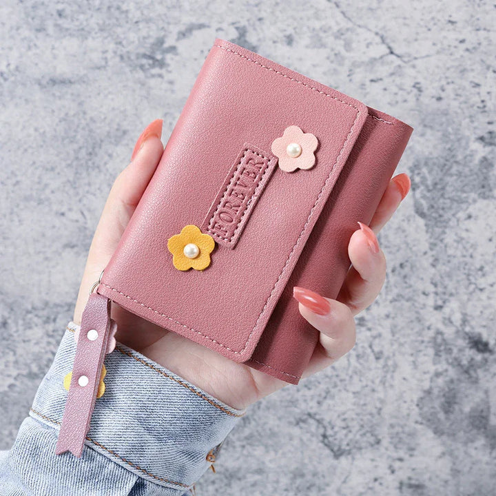 Hand holding a pink leather wallet decorated with yellow and pink flower appliques against a gray textured background