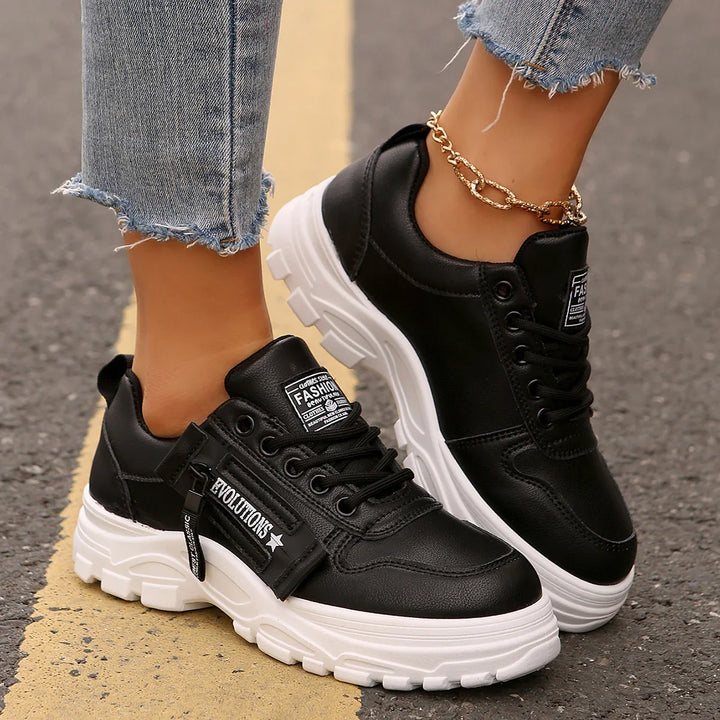 Black chunky sneakers with white thick soles and zipper detail, worn with frayed hem jeans and gold ankle chain