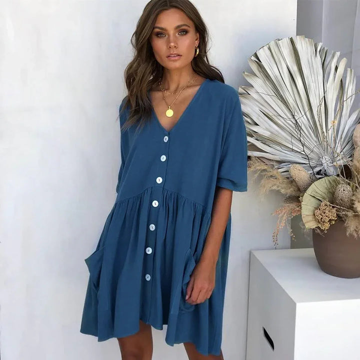 Woman wearing a blue button-down V-neck dress with pockets standing indoors near dried floral decor