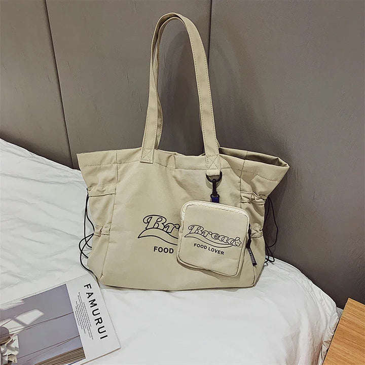 Beige canvas tote bag with matching small pouch, printed with 'Breakfast Food Lover' text, on white bed