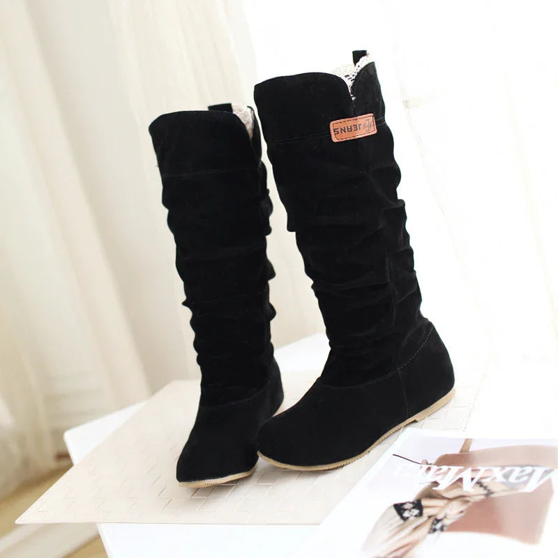 Pair of black suede knee-high boots with flat soles and ruched design displayed on white surface
