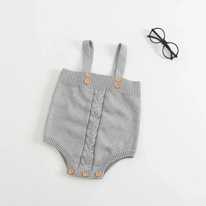 Gray knitted baby romper with straps and wooden buttons laid flat on white background with black round glasses