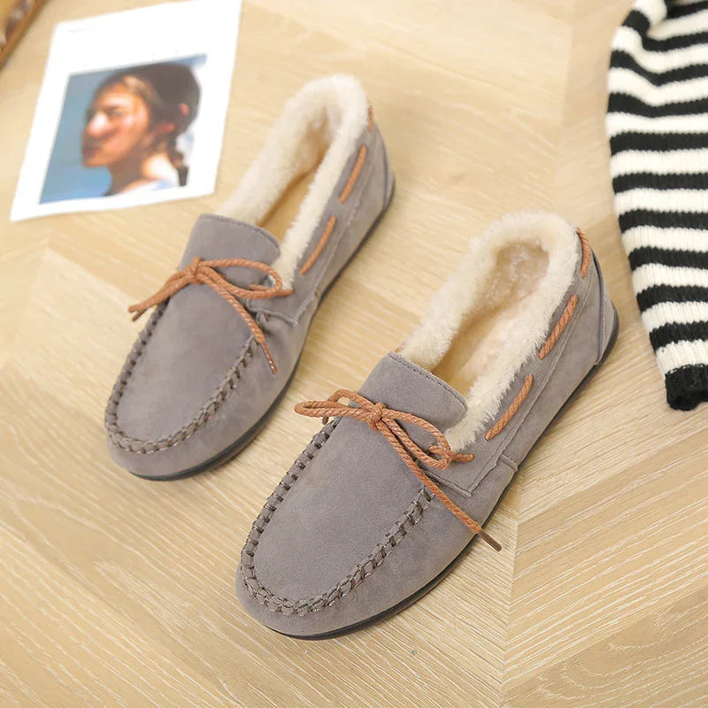 Gray suede moccasin slippers with brown laces and white fur lining on wooden floor