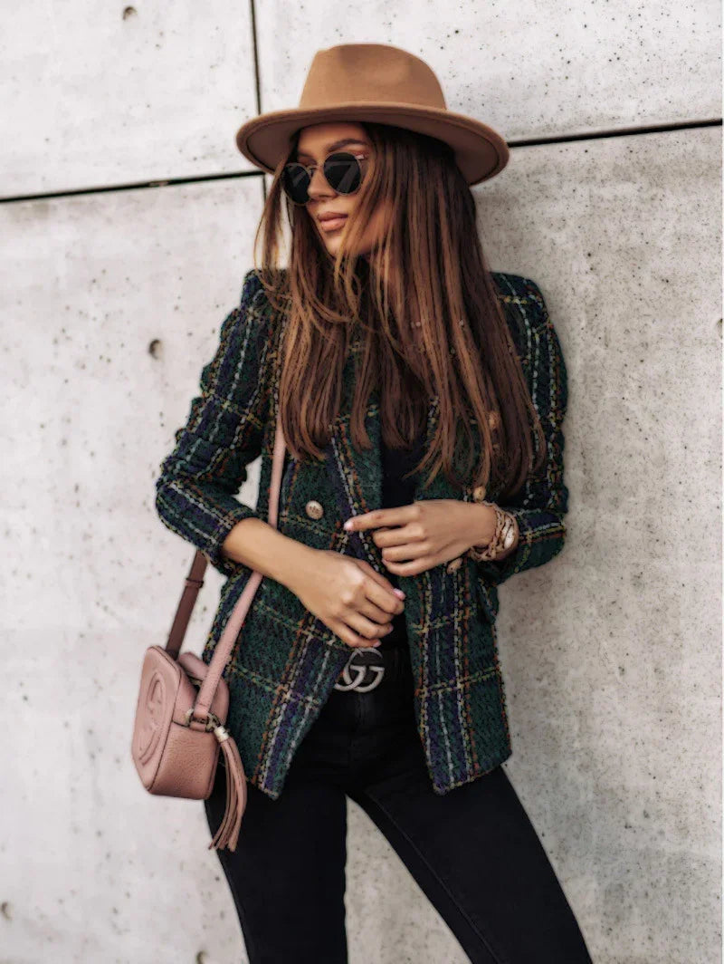 Stylish woman in a plaid blazer, beige hat, black sunglasses, and pink crossbody bag posing against concrete wall