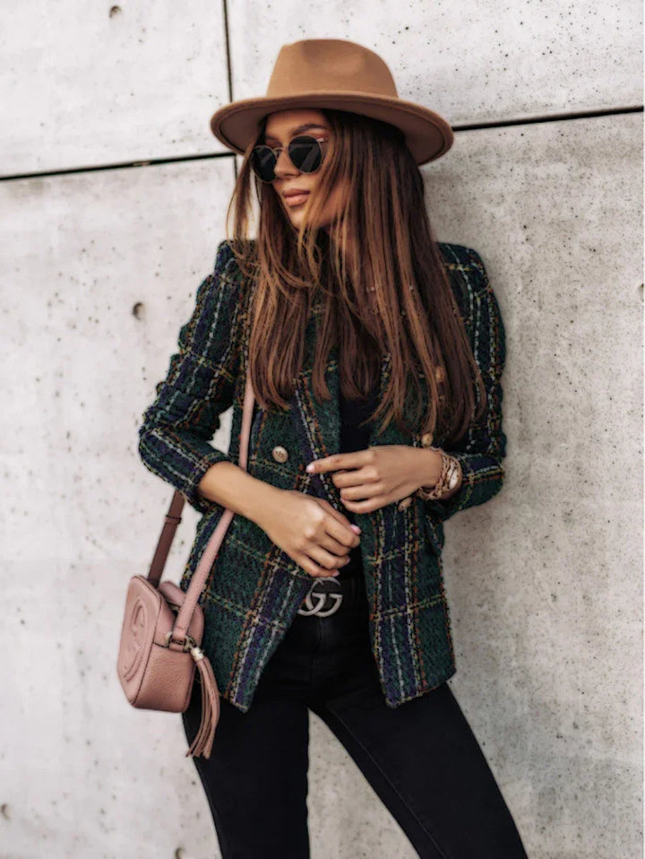 Stylish woman in a plaid blazer, beige hat, black sunglasses, and pink crossbody bag posing against concrete wall