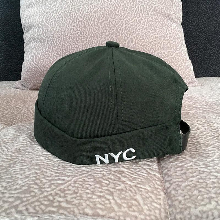 Dark green NYC embroidered beanie cap placed on light textured fabric surface with cushion in background