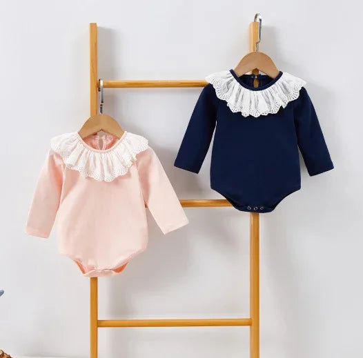 Two baby bodysuits with lace collars, one pink and one navy blue, hanging on wooden ladder