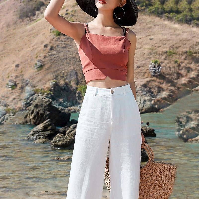 Woman in rust-colored crop top, white wide-leg pants, black sun hat, and hoop earrings by rocky seaside
