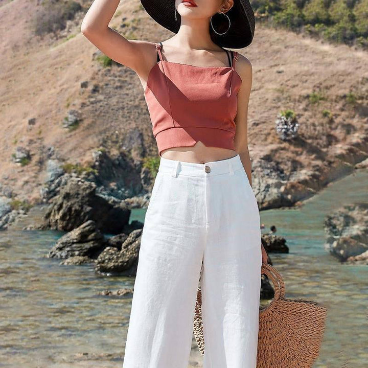 Woman in rust-colored crop top, white wide-leg pants, black sun hat, and hoop earrings by rocky seaside