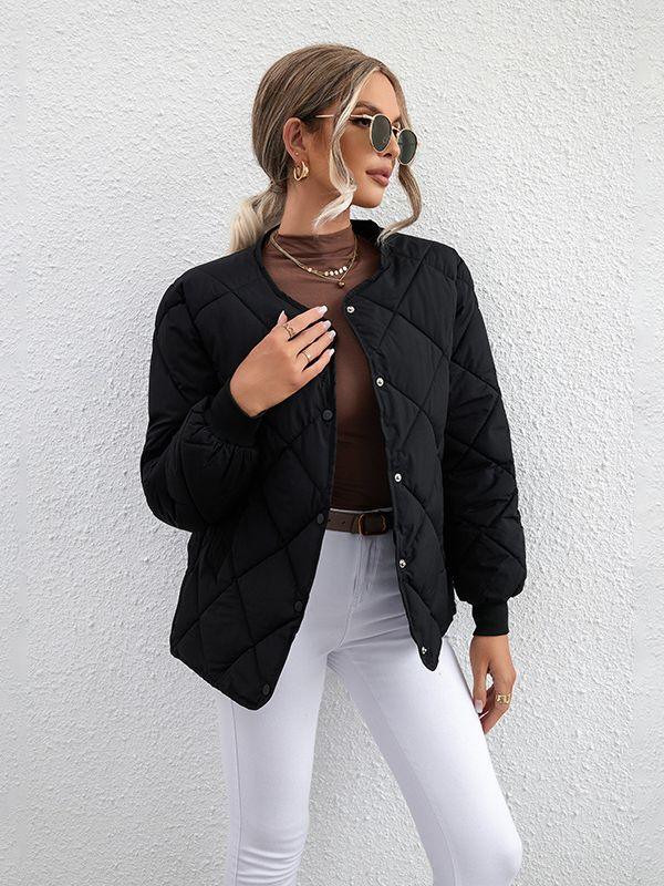 Woman wearing black quilted button-up jacket, brown top, white pants, sunglasses, and gold jewelry against white wall