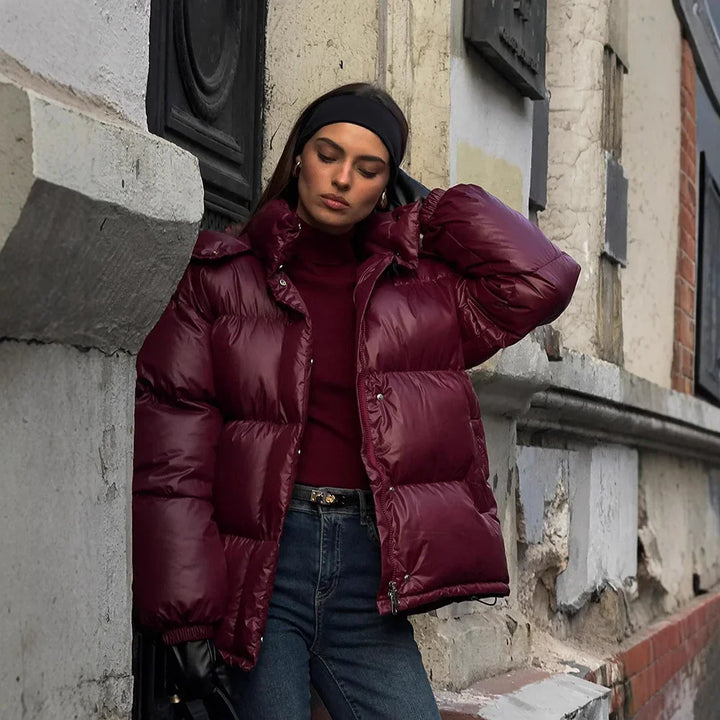 Woman in a shiny burgundy puffer jacket, dark headband, and jeans posing by a weathered urban wall