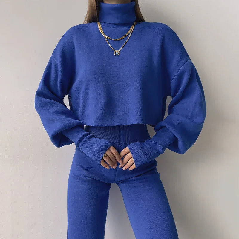 Woman wearing royal blue turtleneck cropped sweater and matching high-waisted pants outfit with gold layered necklace