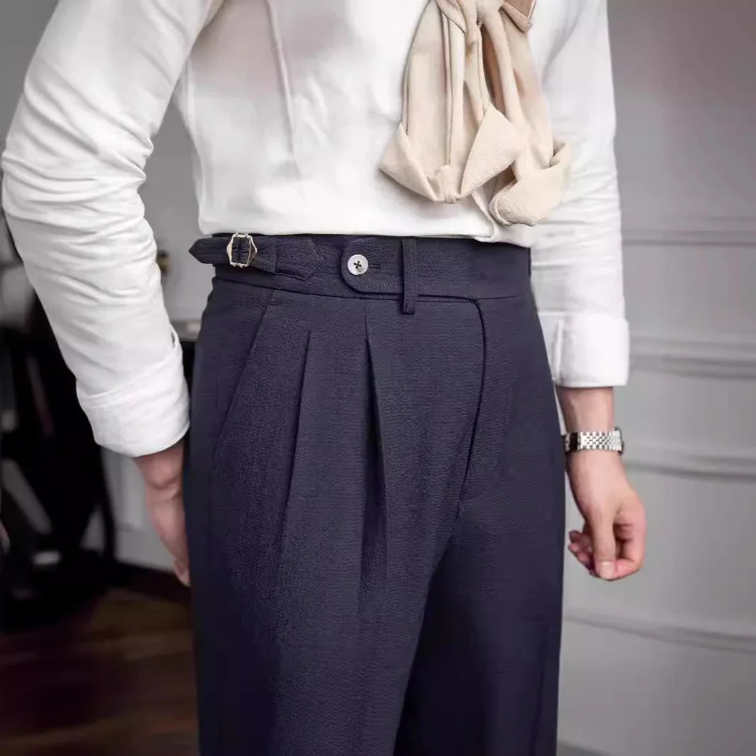 Man wearing navy blue pleated trousers with buckle detail, white shirt, and beige scarf accessory