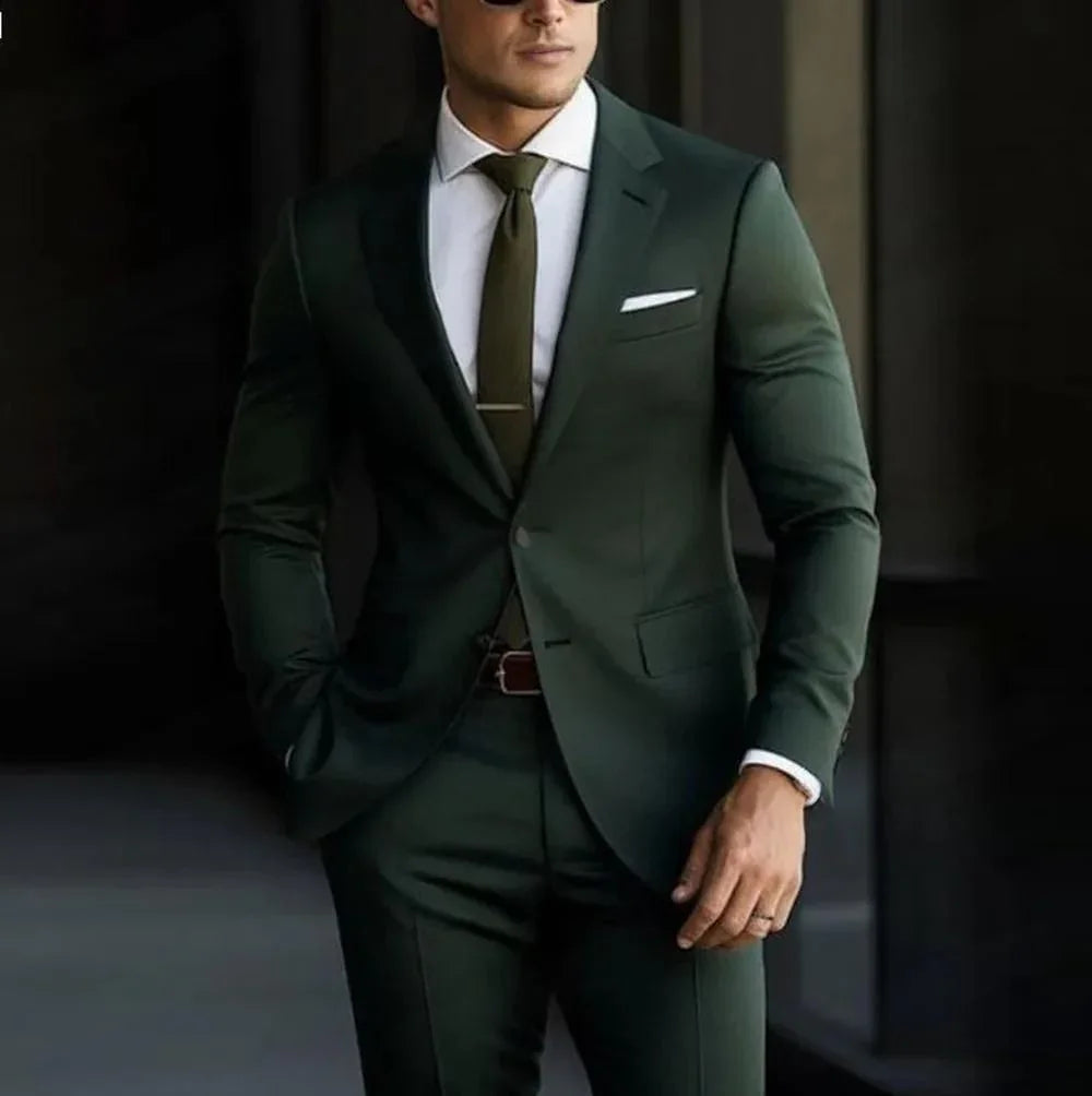 Man wearing a dark green slim-fit suit with white dress shirt, green tie, pocket square, and sunglasses
