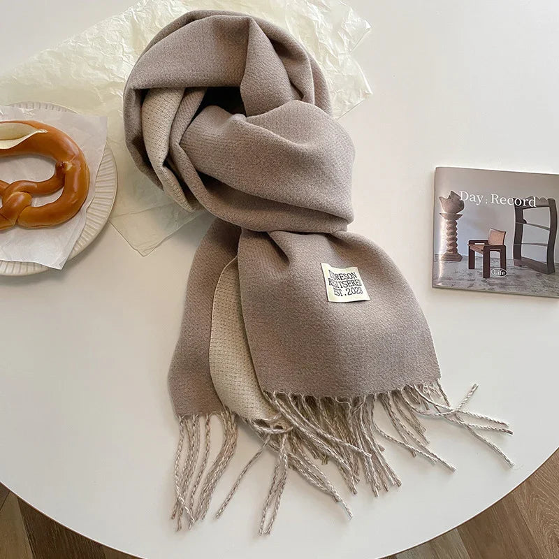 Soft beige wool scarf with fringes on white table next to pretzel and open book